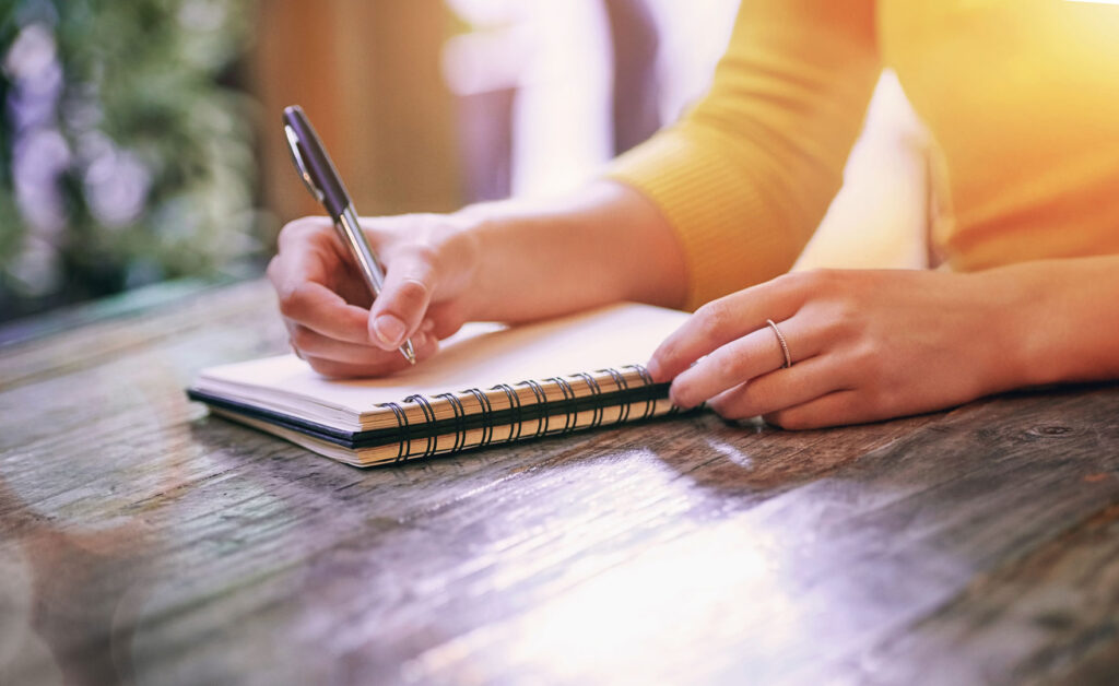 Person writing notes in spiral notebook at Clarity Psychology on rustic wooden table.